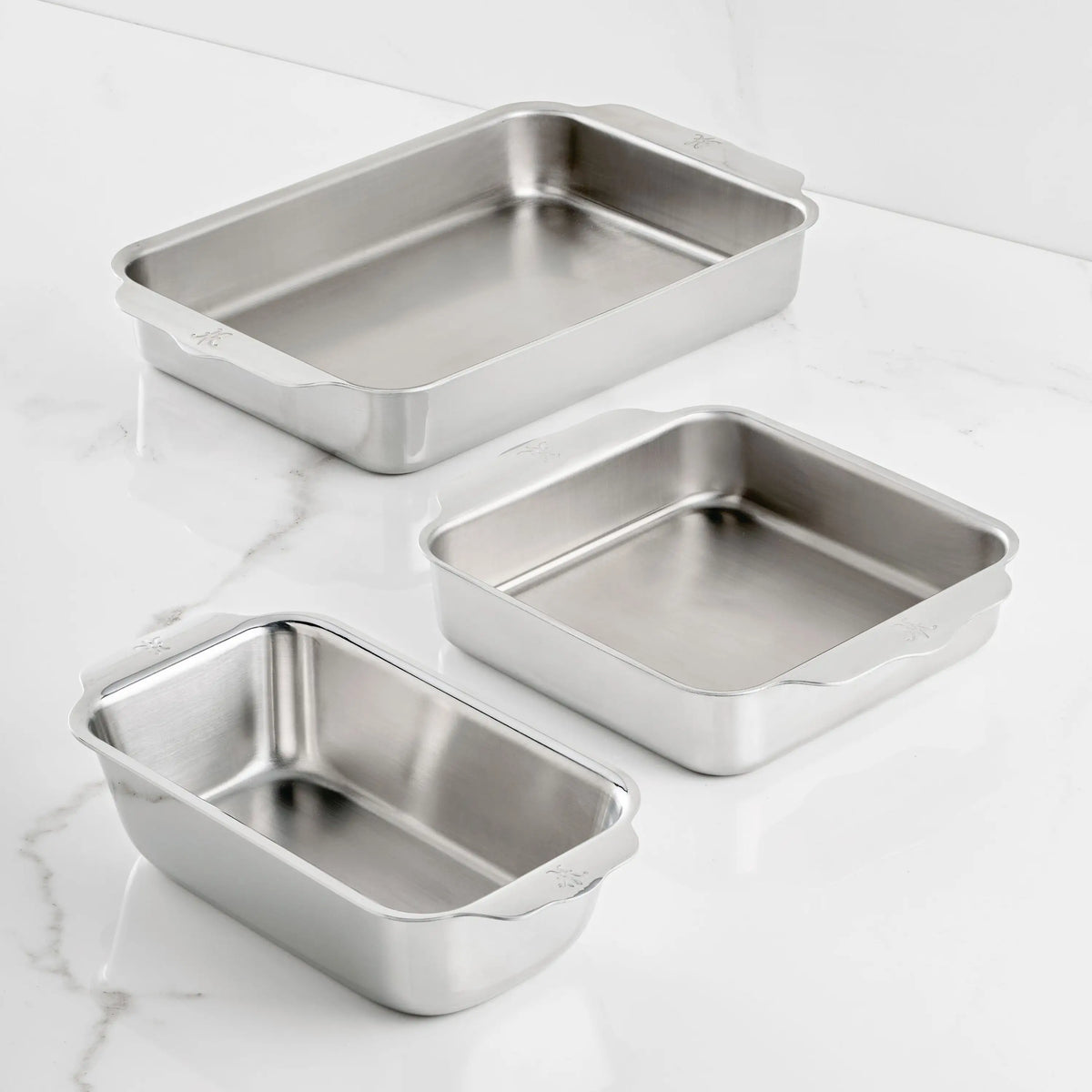 Three empty Hestan OvenBond Tri-Ply 3-Piece Baking Set pans, made of stainless steel with rounded corners and handles, are arranged on a white marble surface. The rectangular pans feature triple-bonded clad construction.