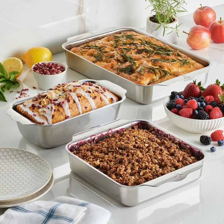 The Hestan OvenBond Tri-Ply 3-Piece Baking Set holds rosemary focaccia, glazed fruit loaf, and berry crumble. Fresh fruit and herbs nearby show this stainless steel set's versatility—an essential for any kitchen.