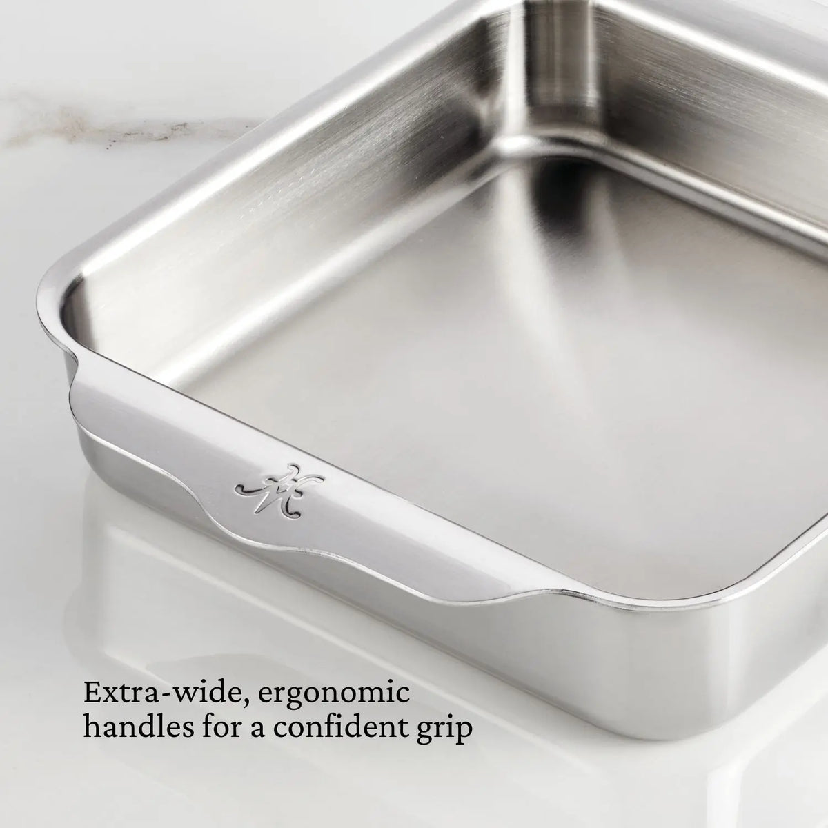 The Hestan OvenBond Tri-Ply 3-Piece Baking Set is displayed on a white marble surface. Extra-wide, ergonomic handles ensure a confident grip, as noted in black text at the bottom left.