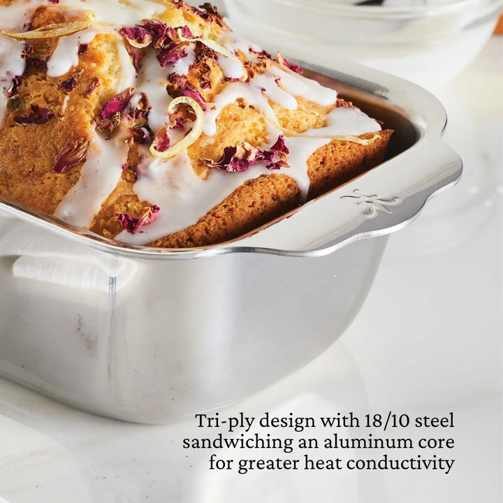 A close-up of a loaf cake with icing and flower petals in a stainless steel loaf pan. Text highlights Hestan OvenBond’s Tri-Ply 18/10 steel with an aluminum core—part of the OvenBond Tri-Ply 3-Piece Baking Set.
