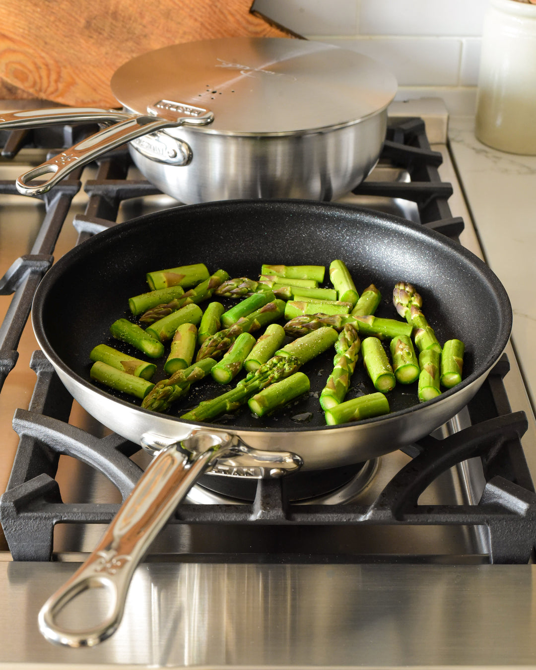 A frying pan with chopped asparagus on a stove. In the background, a covered pot is on another burner. The kitchen surface is white, and there is a small jar beside the stove.