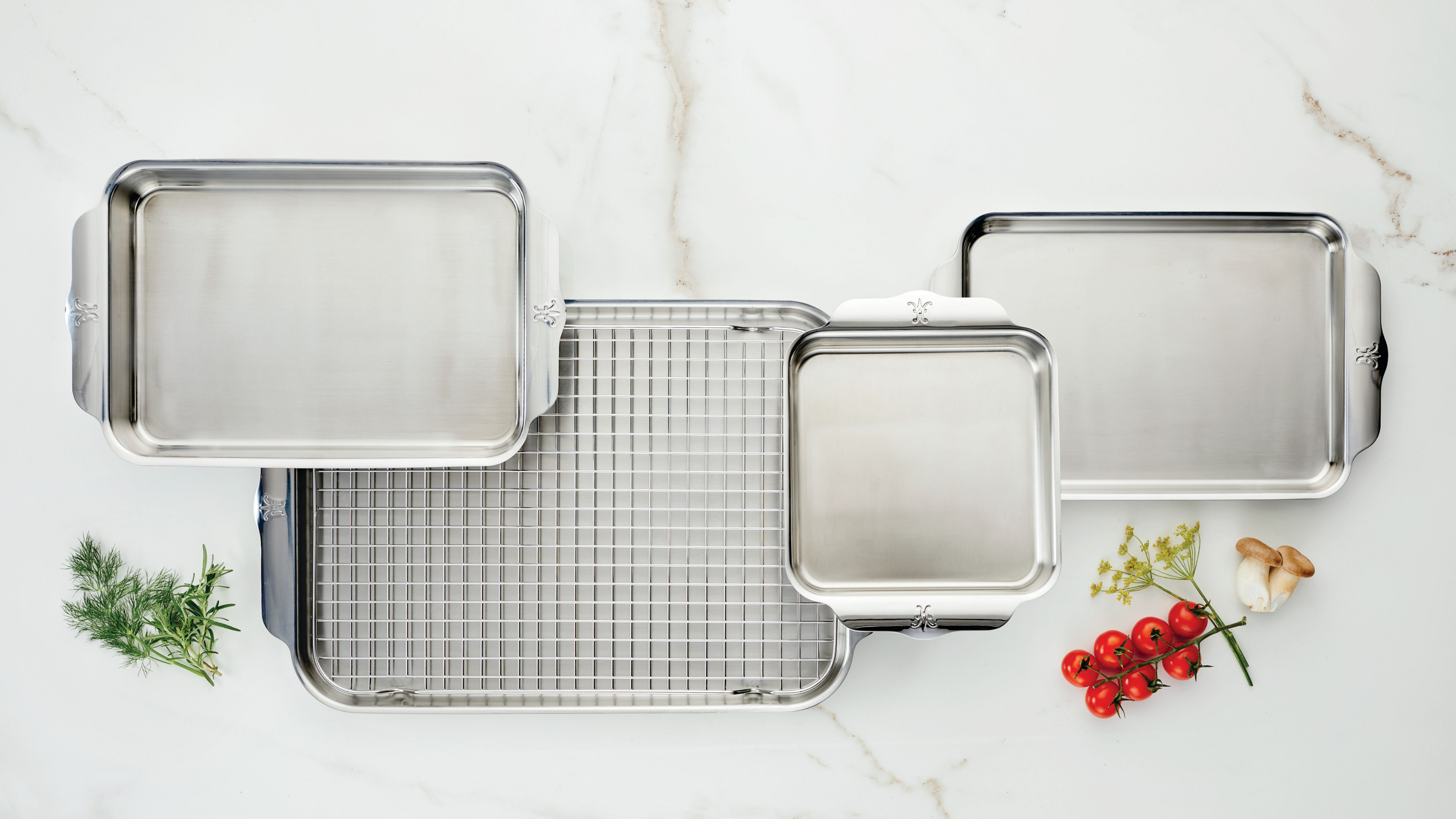 Stainless steel baking trays and a cooling rack are arranged on a white marble surface. Fresh dill, cherry tomatoes on the vine, and mushrooms are placed nearby for garnish.
