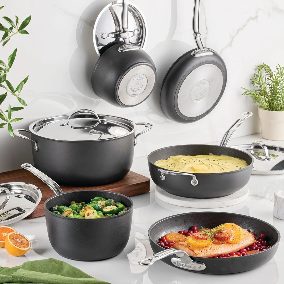 The TITUM® Fusion TITUM Nonstick Cookware Set, 10-piece, in black and silver, is shown on a kitchen counter with food inside. Two pans hang nearby, accented by fresh herbs, a sliced orange, and a white tile backsplash.