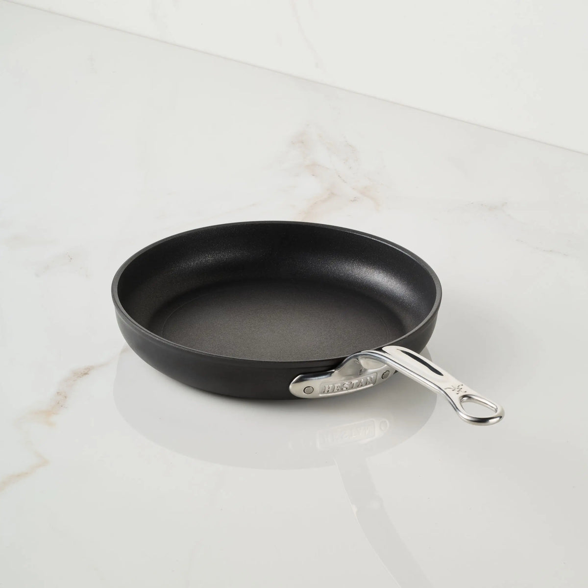 A TITUM® Fusion TITUM Nonstick Open Skillet, featuring a black non-stick surface and silver metal handle, handcrafted in Italy, rests on a white marble surface with subtle brown veining.