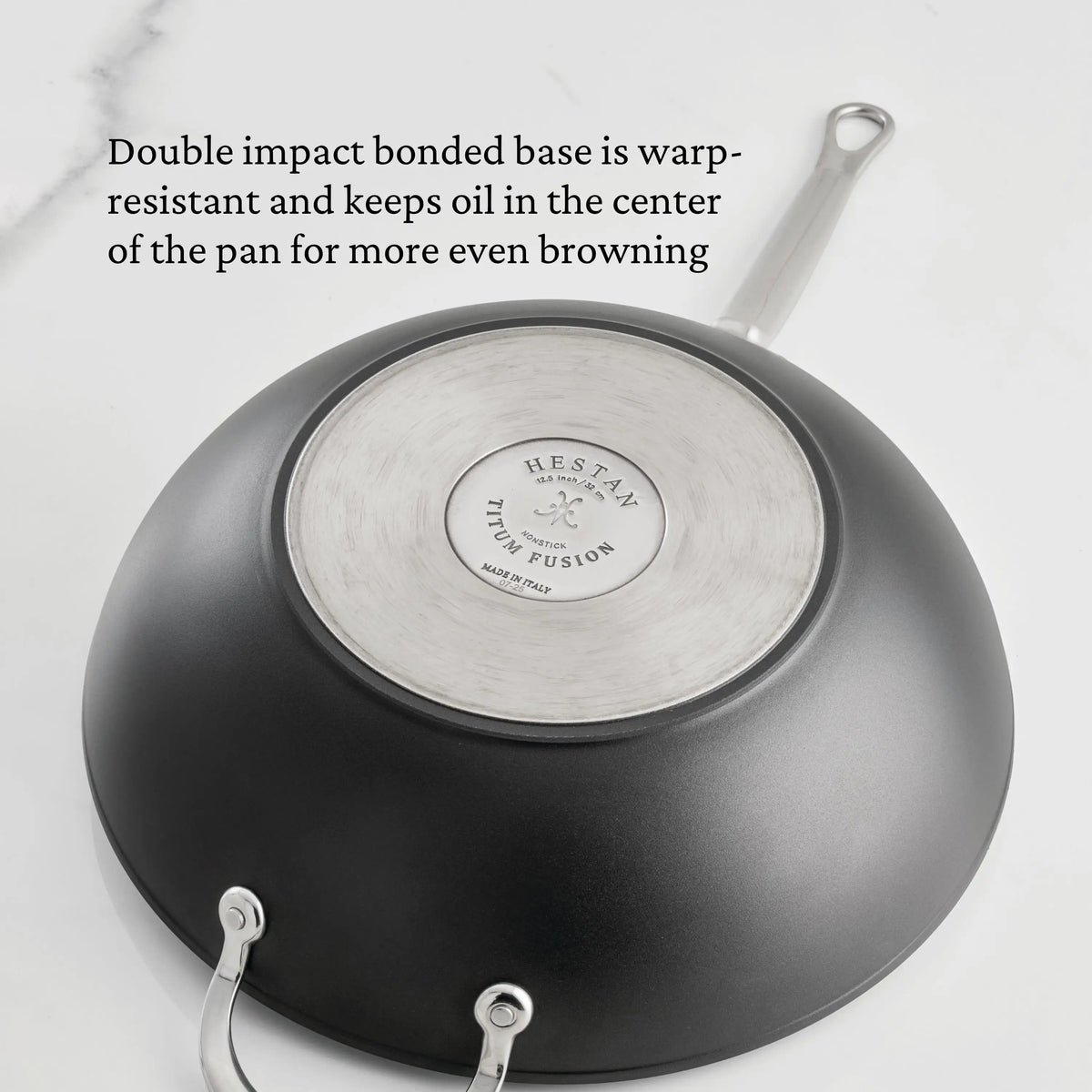 A TITUM® Fusion 12.5-Inch Nonstick Wok with Stick Handle and Dome Lid is shown upside down on a white surface, highlighting its durable brushed stainless steel base and Italian craftsmanship for even browning and warp resistance.