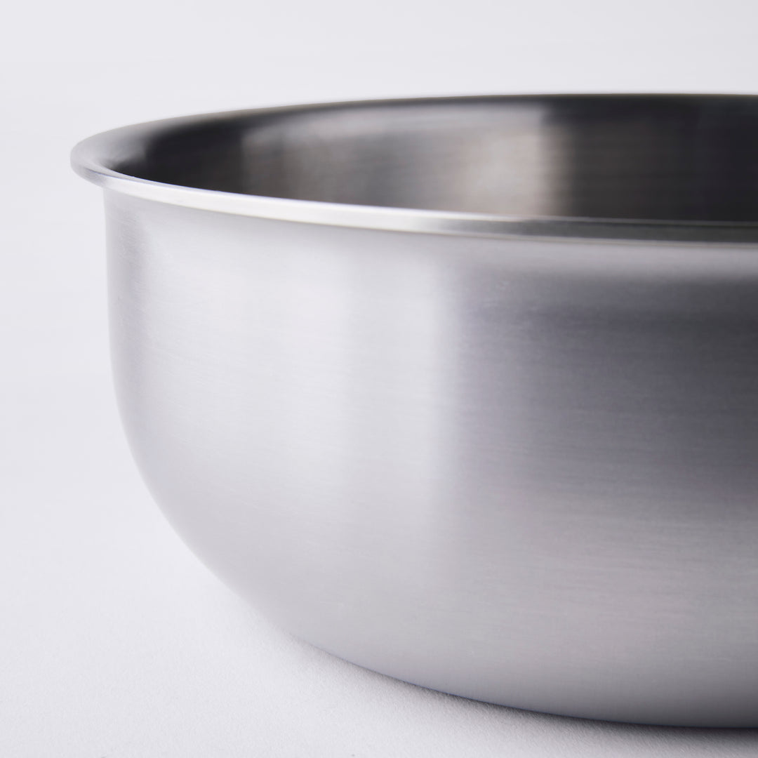 Close-up of a stainless steel mixing bowl on a white background. The focus is on the smooth, reflective surface of the bowl, highlighting its rounded edge and brushed metal texture.