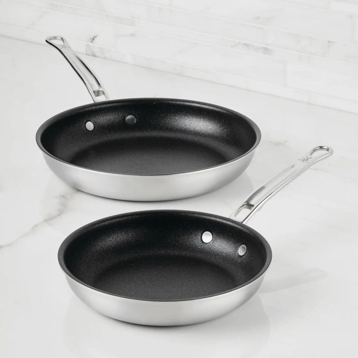 Two Thomas Keller Commercial Clad Stainless Steel TITUM® Nonstick Skillets with black interiors rest on a white marble countertop, offering professional performance and long metal handles—ideal for any kitchen.