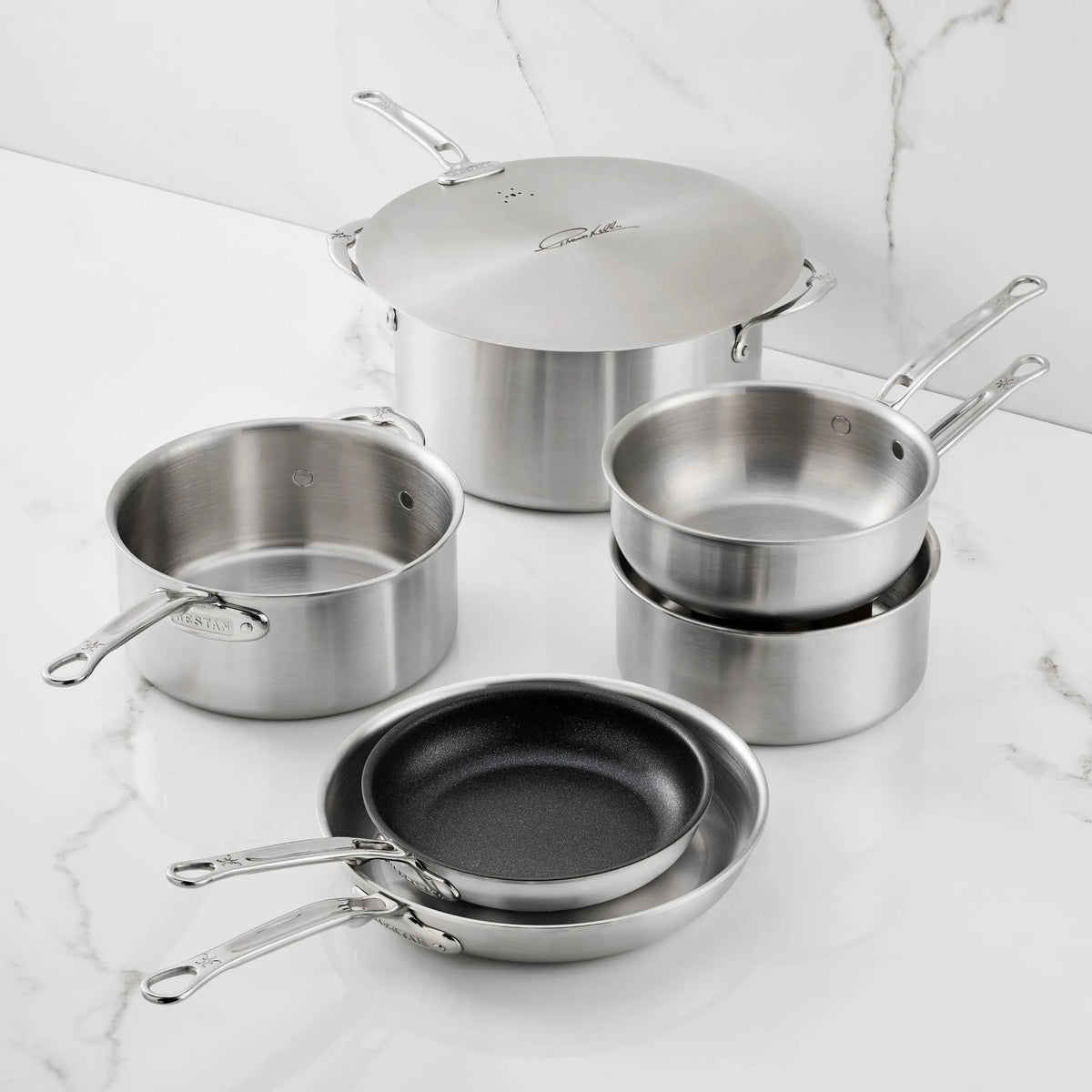 The Thomas Keller Commercial Clad Stainless Steel 7-Piece Cookware Set, featuring saucepans, pots with lids, and nonstick frying pans, is arranged on a white marble countertop for professional culinary performance.