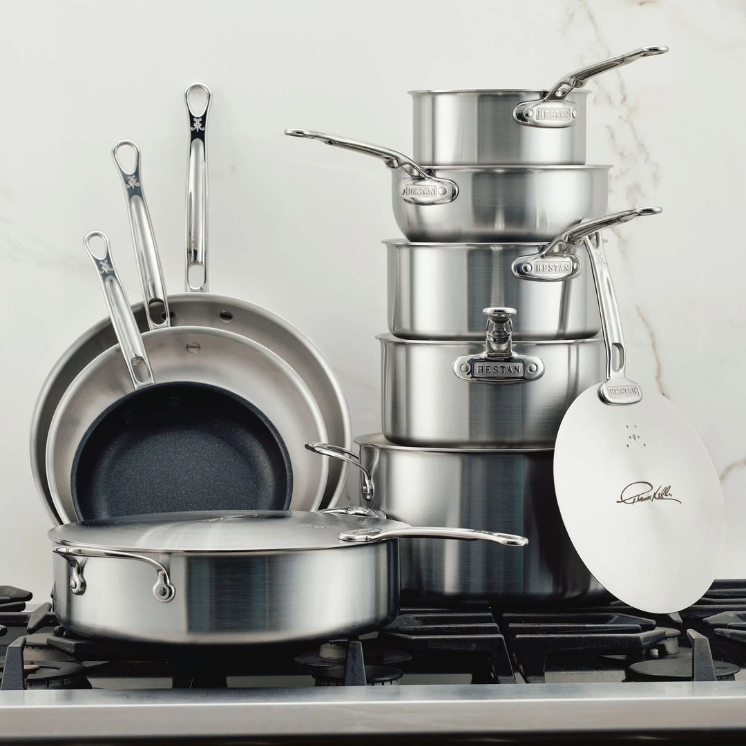 A collection of stainless steel cookware, including pans and pots with lids, is arranged on a stovetop. The set is neatly stacked, showcasing various sizes. Some lids have handles, and the background is a light-colored marble surface.