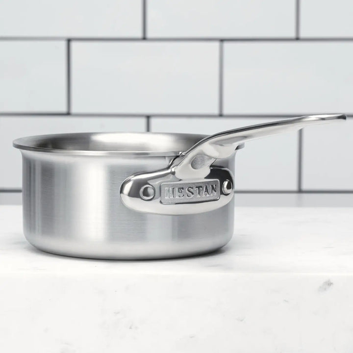 The Thomas Keller Commercial Clad Stainless Steel 0.75-Quart Butter Warmer rests on a white surface before a white tile wall, with the word "Hestan" engraved on its handle attachment.