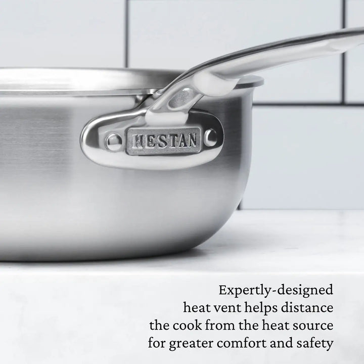Close-up of a Thomas Keller Commercial Clad Stainless Steel 2-Quart Saucier with HEFTAN engraved on the handle, set against tiles. Text below notes its tri-ply clad design for comfort, safety, and professional-grade culinary performance.