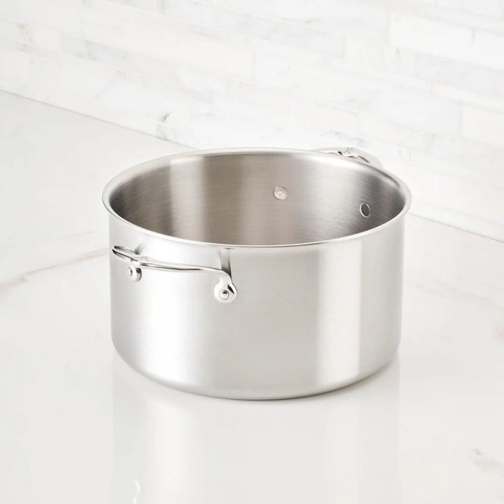 The Thomas Keller Commercial Clad Stainless Steel Stock Pot, with two handles, sits on a white marble countertop before a white tile backsplash, delivering professional-grade performance and sleek style.