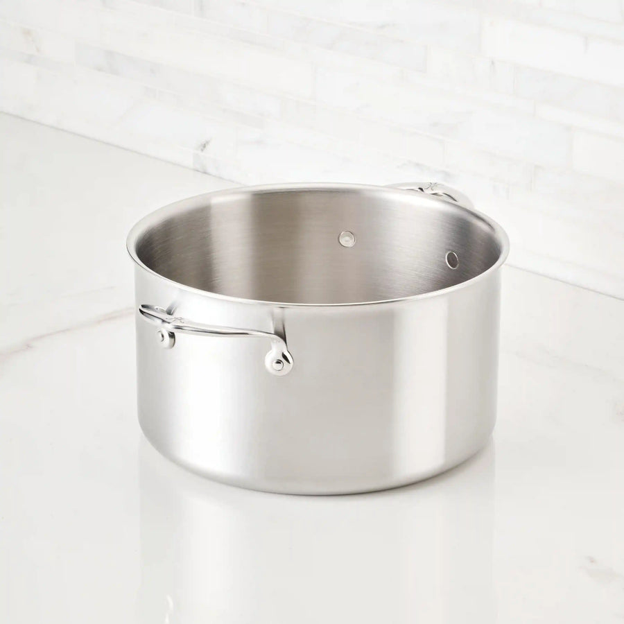 The Thomas Keller Commercial Clad Stainless Steel Stock Pot, with two handles, sits on a white marble countertop before a white tile backsplash, delivering professional-grade performance and sleek style.