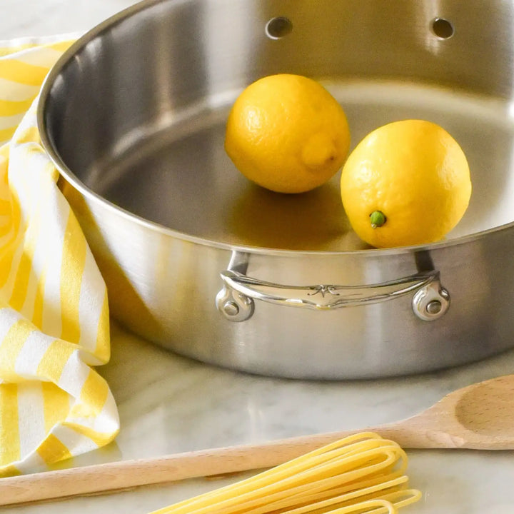 A Thomas Keller Commercial Clad Stainless Steel Rondeau with two whole lemons inside rests next to a yellow and white striped kitchen towel, a wooden spoon, and uncooked spaghetti on a marble countertop—perfect cookware for any aspiring chef.