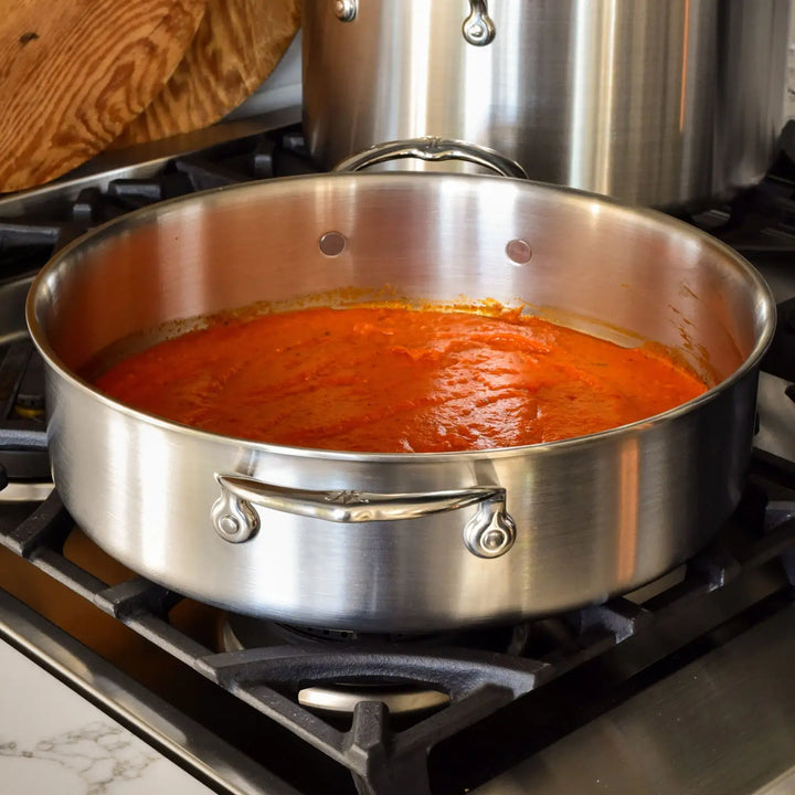 A Thomas Keller Commercial Clad Stainless Steel Rondeau filled with red tomato sauce sits on a stovetop burner, with a wooden cutting board and another pot visible in the background.
