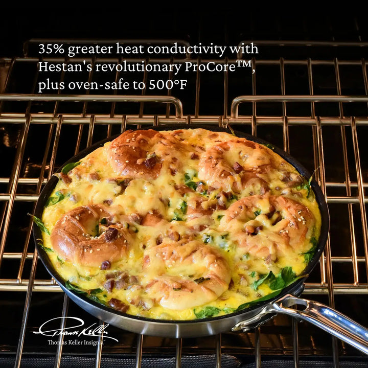 A Thomas Keller Commercial Clad Stainless Steel TITUM® Nonstick Skillet with a baked egg and bread dish sits on an oven rack. Text notes 35% more heat conductivity, oven-safe up to 500°F, and pro performance. A signature appears at the lower left.