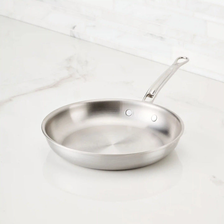 A Thomas Keller Commercial Clad Stainless Steel Skillet with a long, riveted handle sits clean and empty on a white marble countertop against a light tiled backsplash.