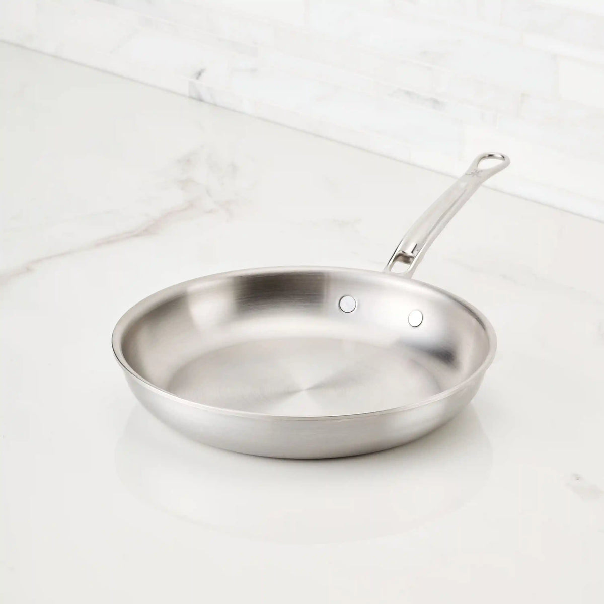 A Thomas Keller Commercial Clad Stainless Steel Skillet with a long, riveted handle sits clean and empty on a white marble countertop against a light tiled backsplash.