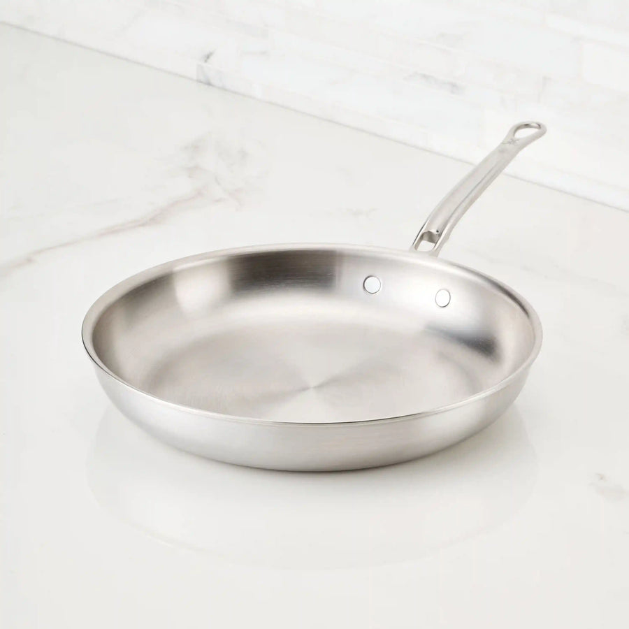 A Thomas Keller Commercial Clad Stainless Steel Skillet with a metal handle sits on a white marble countertop in a bright kitchen, showcasing the professional quality of Stainless Steel Skillets.