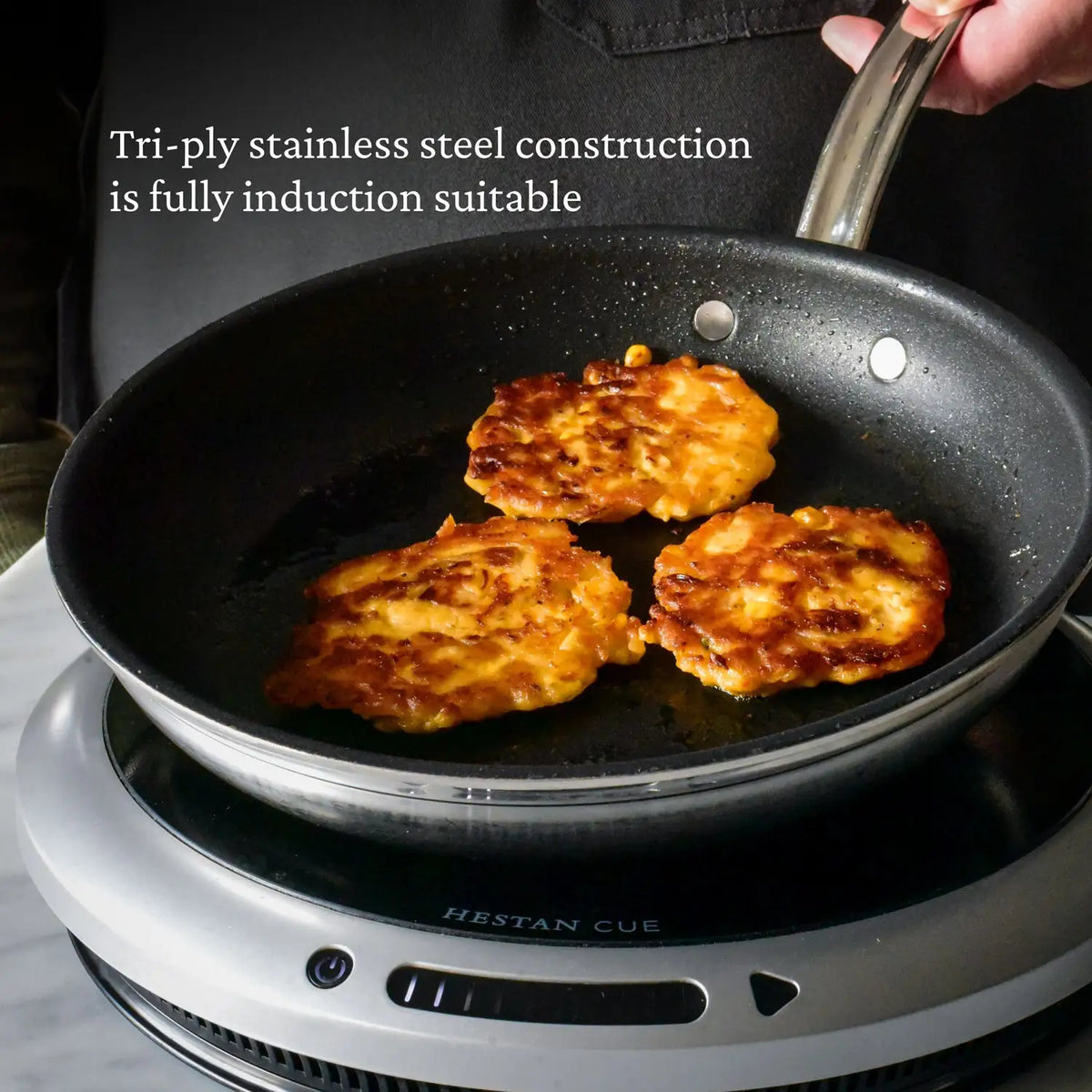 A ProBond® Forged™ Professional Clad Stainless Steel TITUM® Nonstick Skillet with three browned fritters cooks on an induction cooktop. Text reads, Tri-ply stainless steel construction is fully induction suitable.