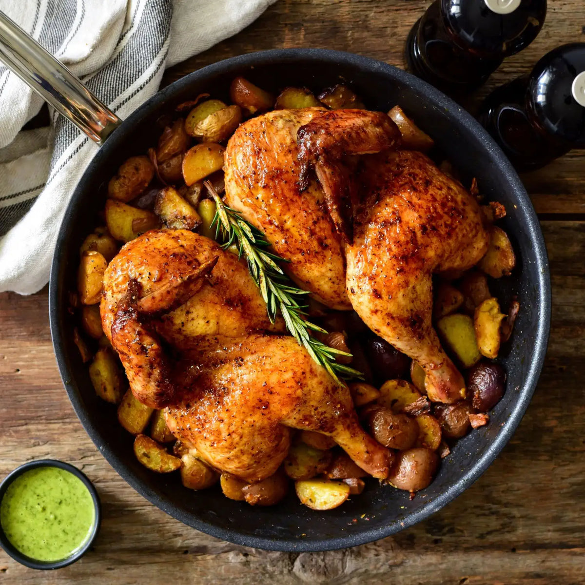 A ProBond® Forged™ Brushed Clad Stainless Steel TITUM® Nonstick Skillet holds two roasted spatchcocked chickens on mixed potatoes with rosemary. A cloth napkin, salt and pepper shakers, and a bowl of green sauce are nearby on a wooden surface.