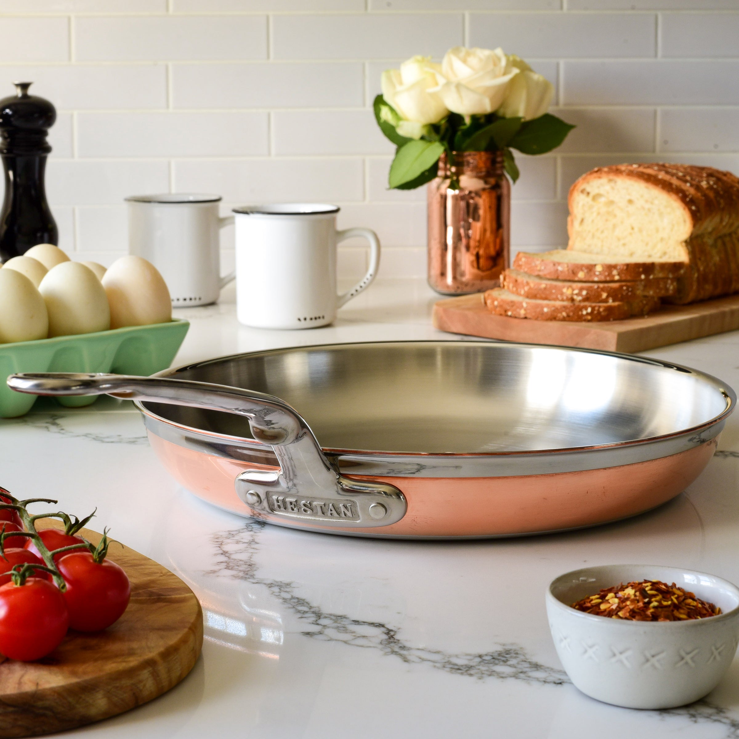 A copper frying pan with a stainless steel interior is on a kitchen counter. Nearby are a loaf of bread, a bowl of red pepper flakes, cherry tomatoes, eggs in a carton, two mugs, a pepper grinder, and a vase of white roses.