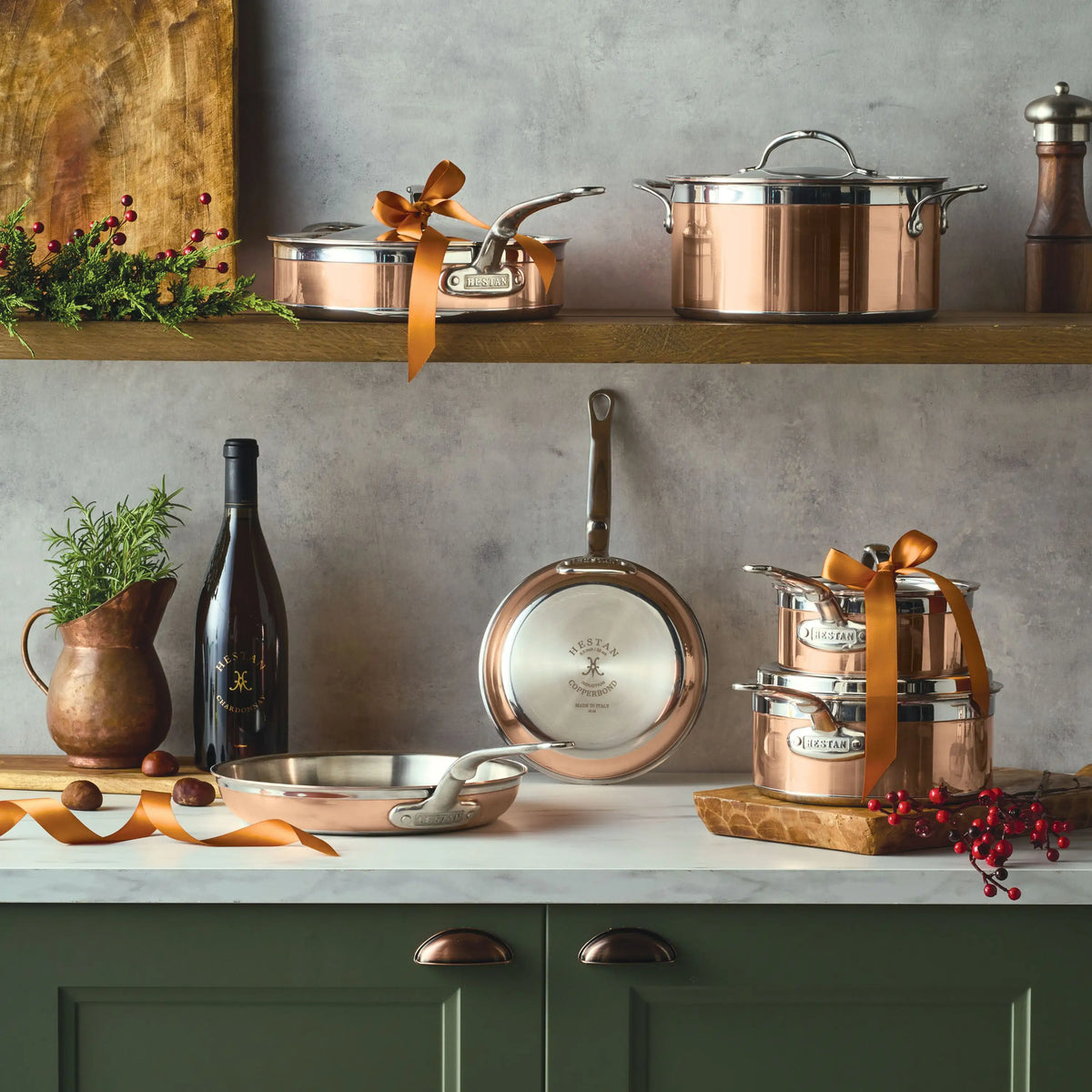 CopperBond® Copper Induction Ultimate Set, 10-Piece cookware is arranged on a countertop and wooden shelf alongside a wine bottle, wooden pitcher, spices, a plant, and decorative berries, all set against a gray wall.