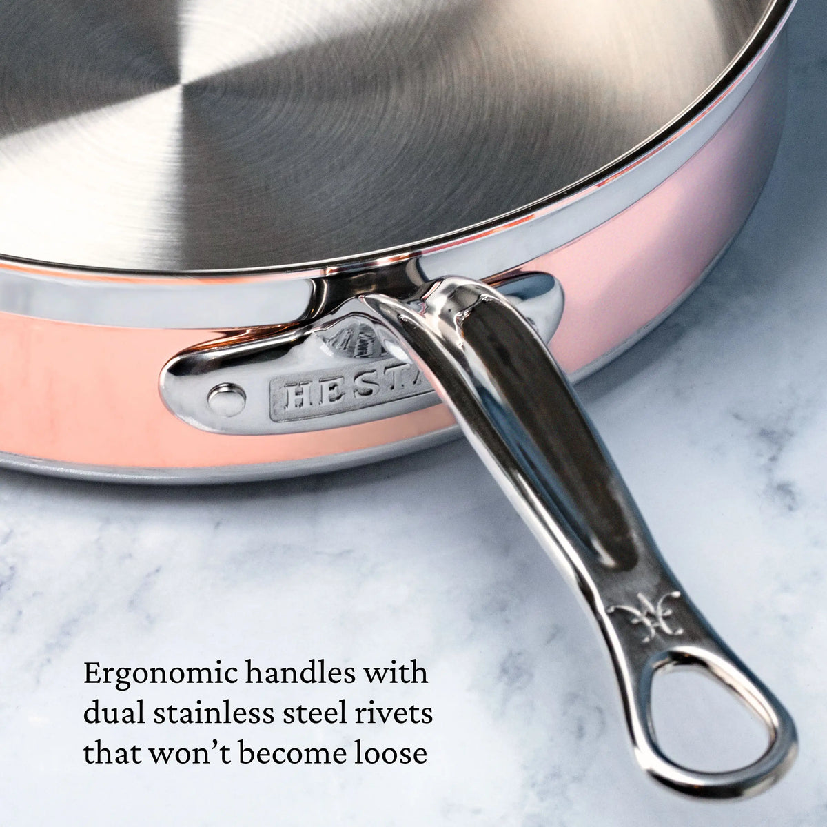 A close-up of a CopperBond Copper Induction Set frying pan, featuring a shiny ergonomic handle secured with dual stainless steel rivets for lasting stability, sits on marble. Text: Ergonomic handles with dual stainless steel rivets that won’t become loose.