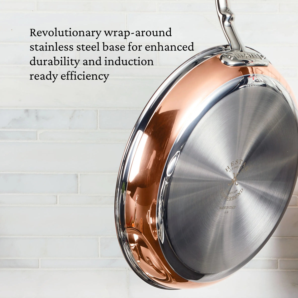 A CopperBond Copper Induction Set frying pan hangs on a white tile wall, featuring a wrap-around stainless steel base for durability and induction-ready efficiency, highlighting the 24-piece set’s premium craftsmanship.