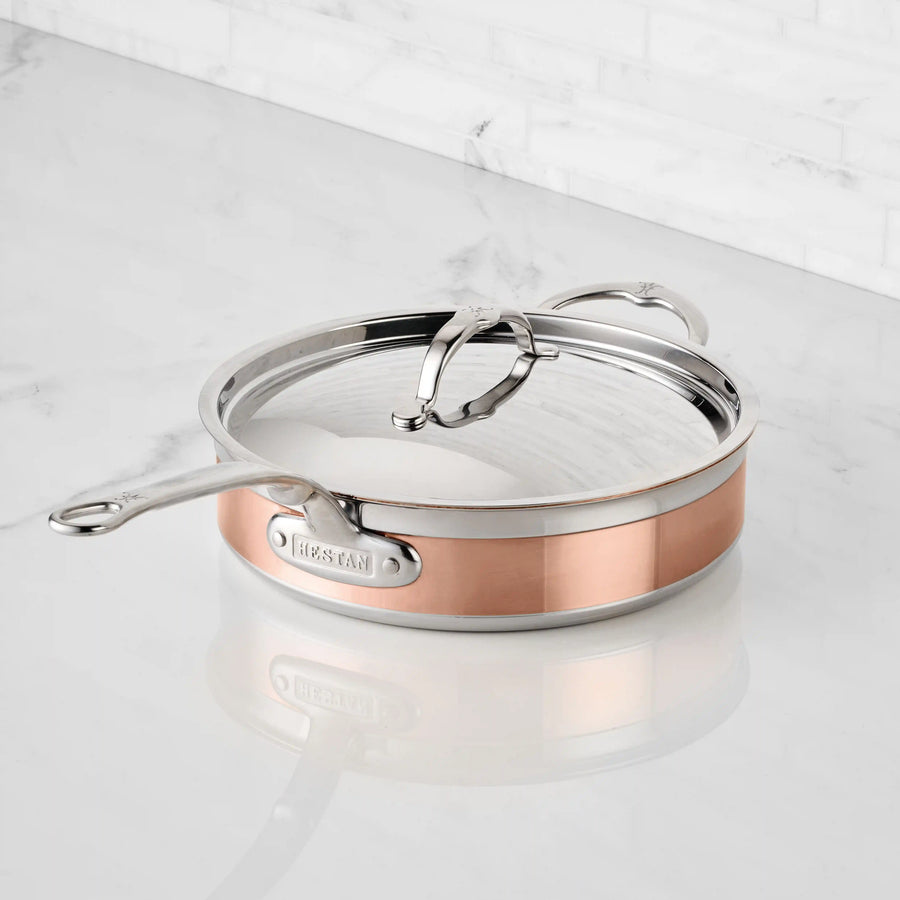 A CopperBond Copper Induction Sauté with Lid, 3.5-Quart, featuring a polished finish and dual handles, sits on a white marble countertop.