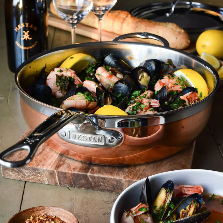The CopperBond Copper Induction Essential Pan, handcrafted in Italy, holds a medley of shrimp and mussels with herbs and lemon wedges. A baguette complements two wine glasses and a bottle, while a small bowl of seafood delights sits in the foreground.