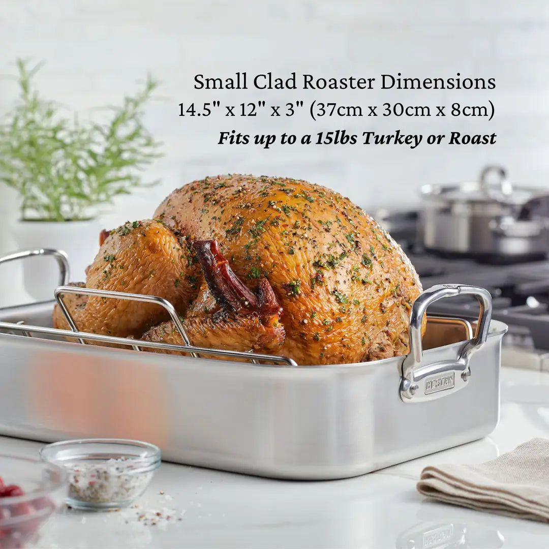 A roasted turkey adorned with herbs sits elegantly in the Hestan Provisions 14.5-inch Classic Clad Roaster with Rack on a kitchen countertop, framed by blurred cookware and a green plant. This stainless steel roasters dimensions are 14.5 x 12 x 3, ideal for roasting up to a 15 lb feast.