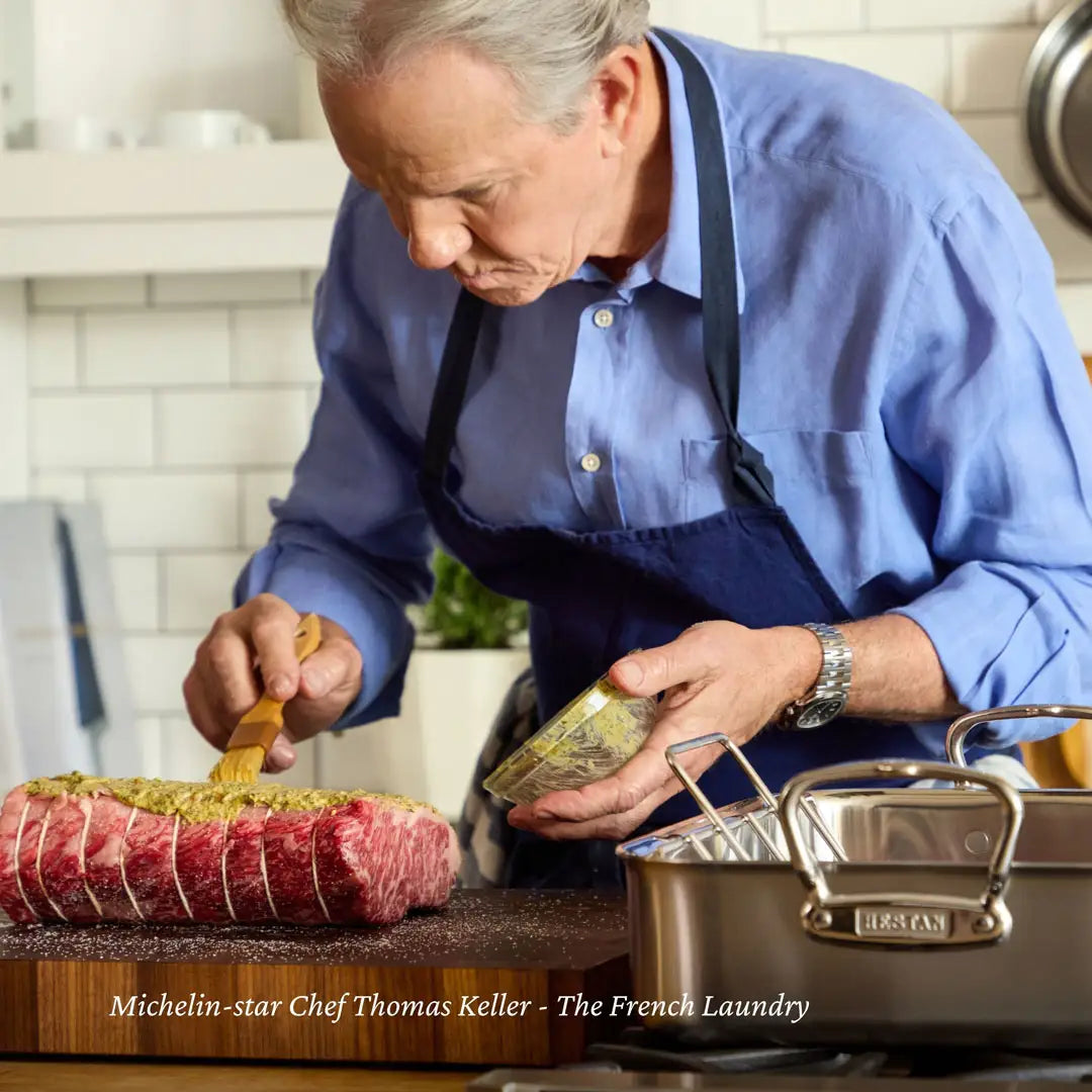 A person in a blue shirt and apron brushes a mixture on tied raw meat in a Hestan Provisions 16.5-inch Classic Clad Roaster with Rack. A pot is seen on the counter. The text reads, Michelin-star Chef Thomas Keller - The French Laundry.