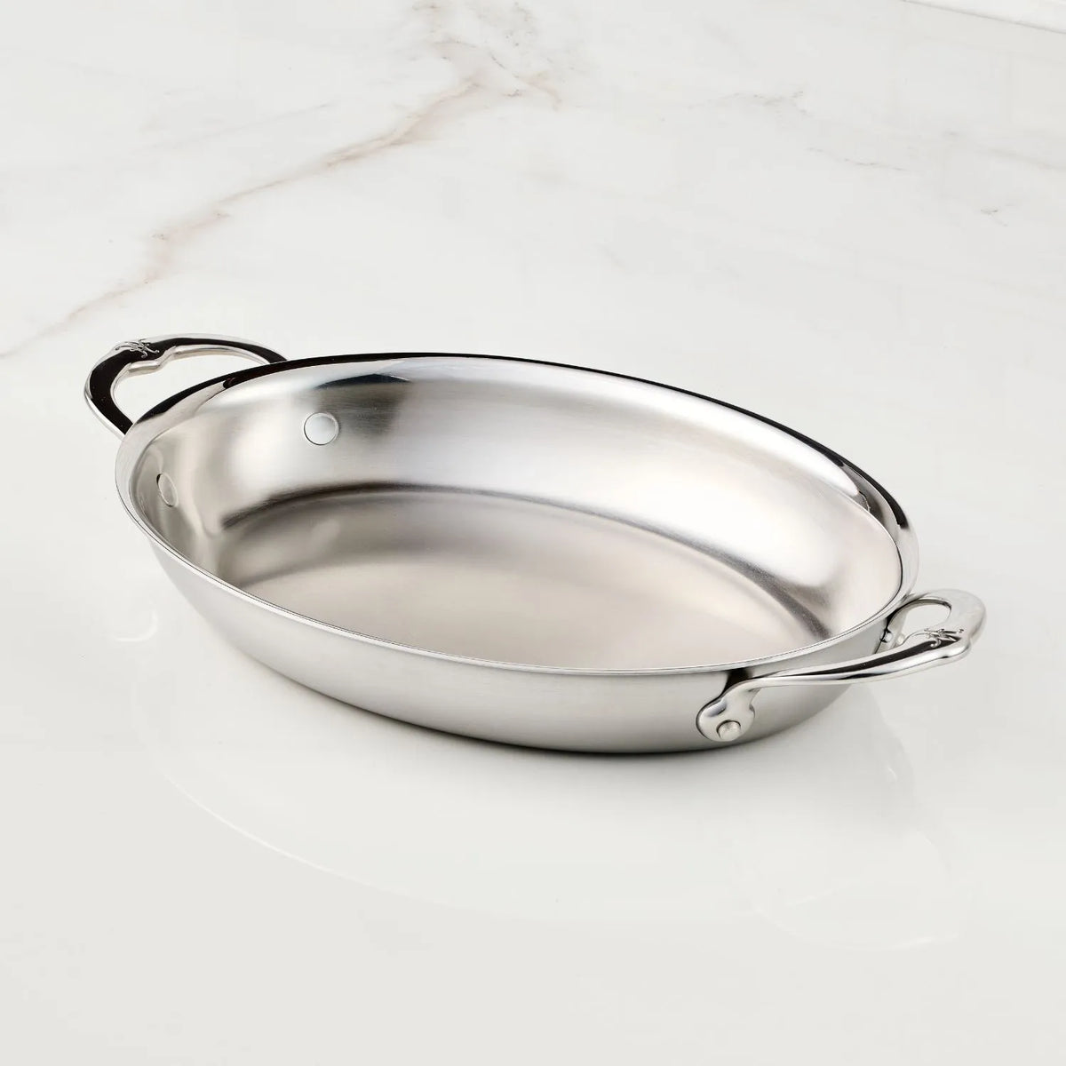 The Hestan Provisions 12.5-Inch x 9-Inch Oval Au Gratin pan, with two handles and a polished stainless steel finish, sits on a white marble surface, highlighting its elegant and seamless design.