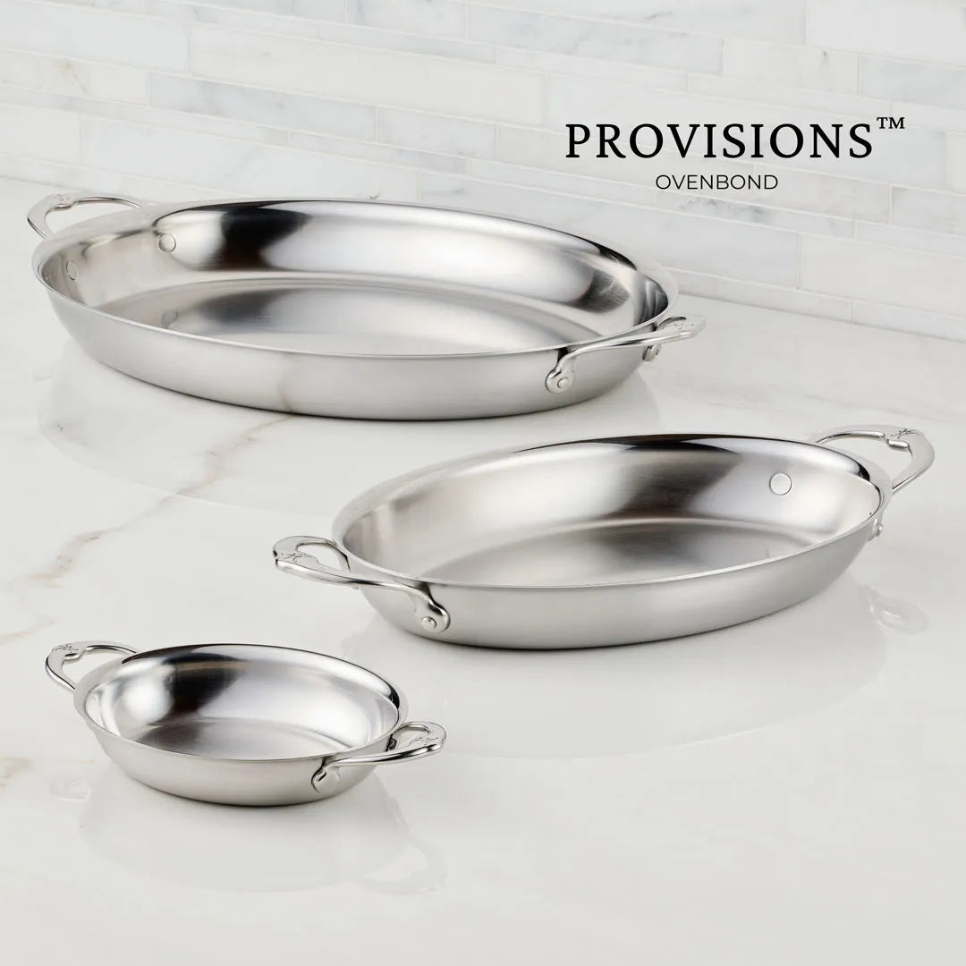 The Hestan Provisions 15-Inch x 9-Inch Oval Au Gratin dish with handles is displayed on a white marble surface against a tiled wall, with the Hestan Provisions Au Gratin logo in the background.