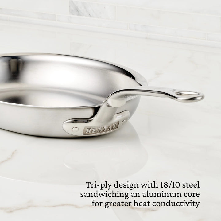 The Hestan Provisions Oval Fish Pan features a silver finish and tri-ply design, with an engraved handle. Corner text highlights its 18/10 stainless steel construction and aluminum core for even heat distribution, resting on a white marble surface.