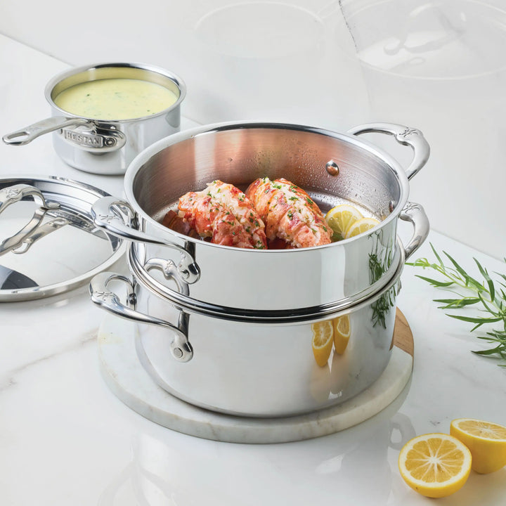 The ProBond® Luxe™ Polished Clad Stainless Steel 3-quart Soup Pot and Steamer Set holds two lobster tails with lemon slices, while a saucepan with yellow sauce, lemon halves, rosemary, and thyme sit on a white countertop.