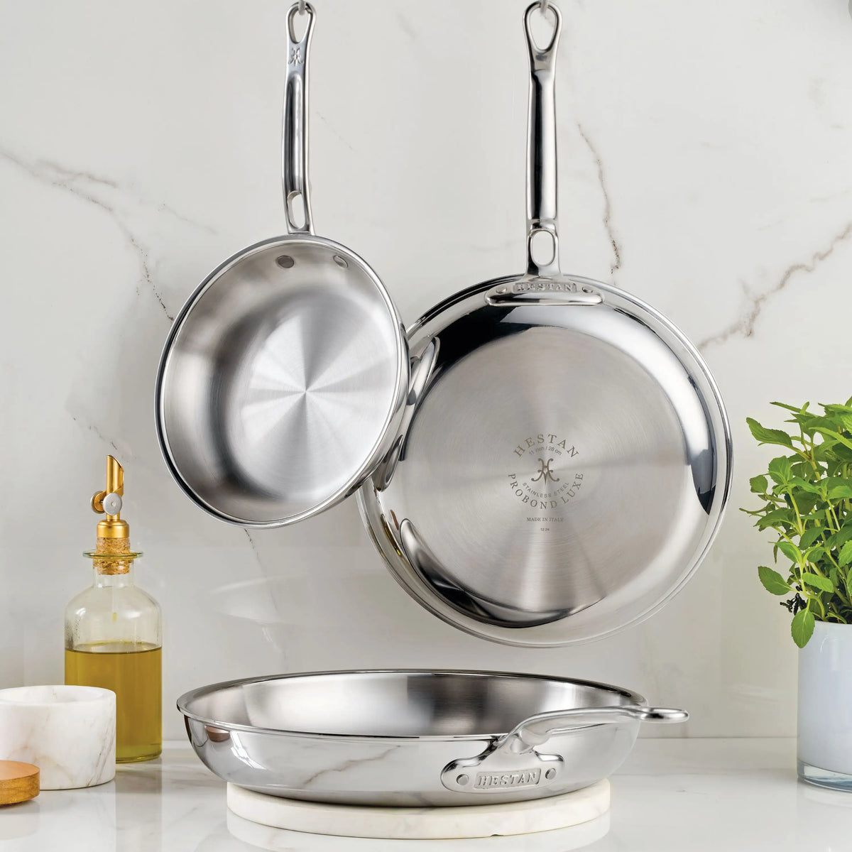 Three Polished Clad Stainless Steel Skillets from ProBond® Luxe™ are shown: two hang on a marble wall, while one rests on the counter next to an olive oil dispenser, marble container, and a green herb plant.