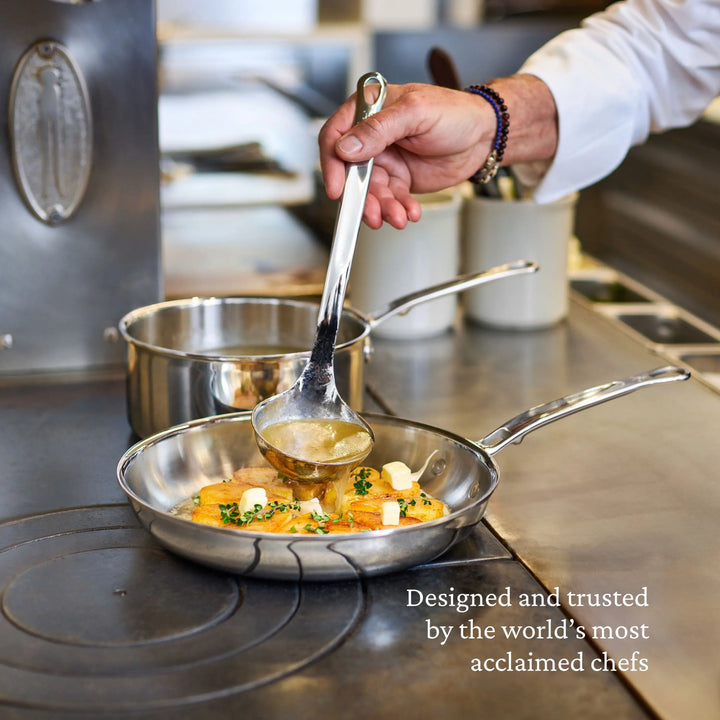 A hand pours liquid with a ladle into a ProBond® Luxe™ Polished Clad Stainless Steel Saucepan with Lid, cooking fish and vegetables on the stove. Text: “Designed and trusted by the world’s most acclaimed chefs.”.
