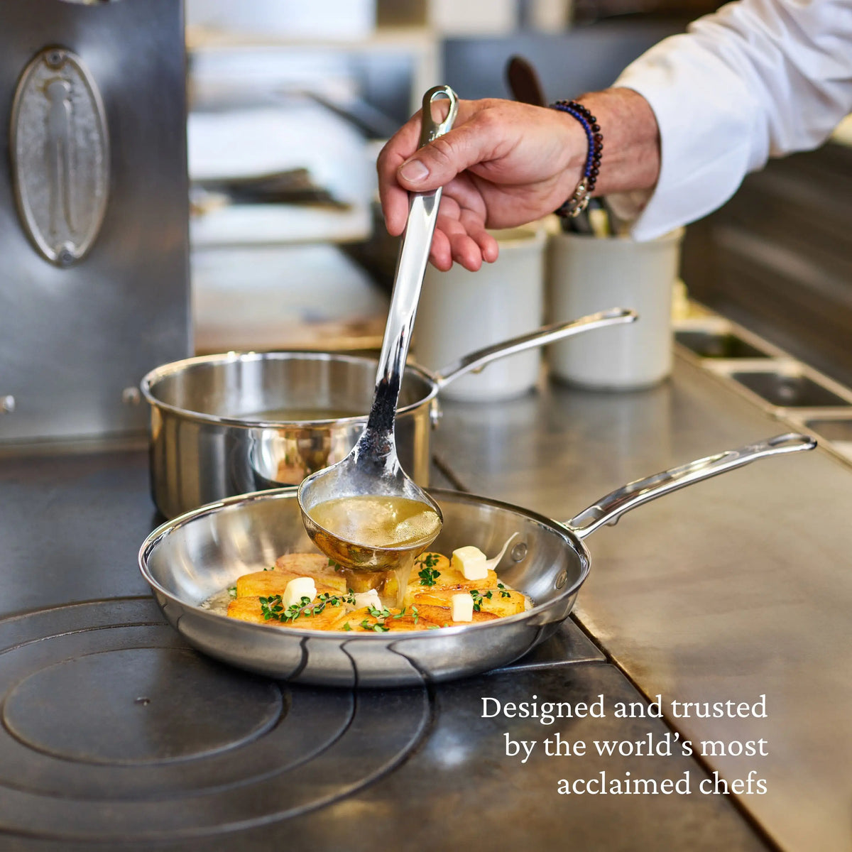 A hand pours liquid with a ladle into a ProBond® Luxe™ Polished Clad Stainless Steel Saucepan with Lid, cooking fish and vegetables on the stove. Text: “Designed and trusted by the world’s most acclaimed chefs.”.