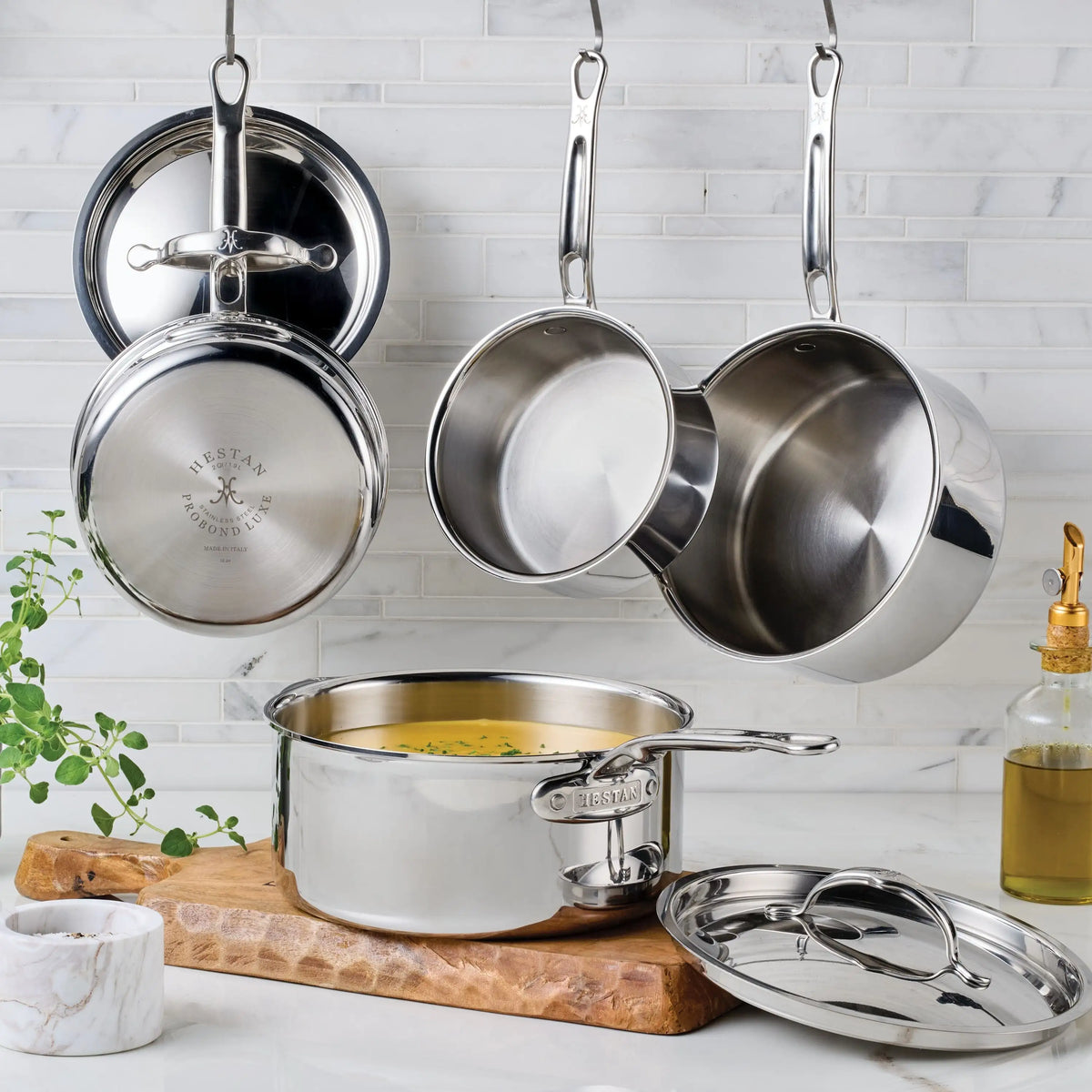 ProBond® Luxe™ Polished Clad Stainless Steel Saucepans with Lids are displayed, some hanging on hooks and others on a wooden board. One saucepan holds soup, alongside a marble bowl, plant, and oil bottle on a white marble countertop.