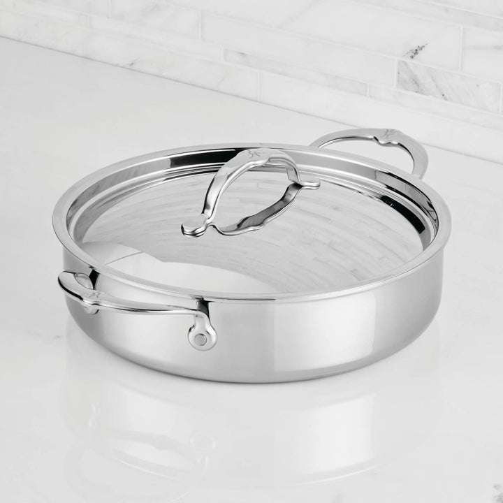 A ProBond® Luxe™ Polished Clad Stainless Steel Rondeau with lid and dual handles rests on a white marble countertop, set against a backdrop of white tile backsplash.