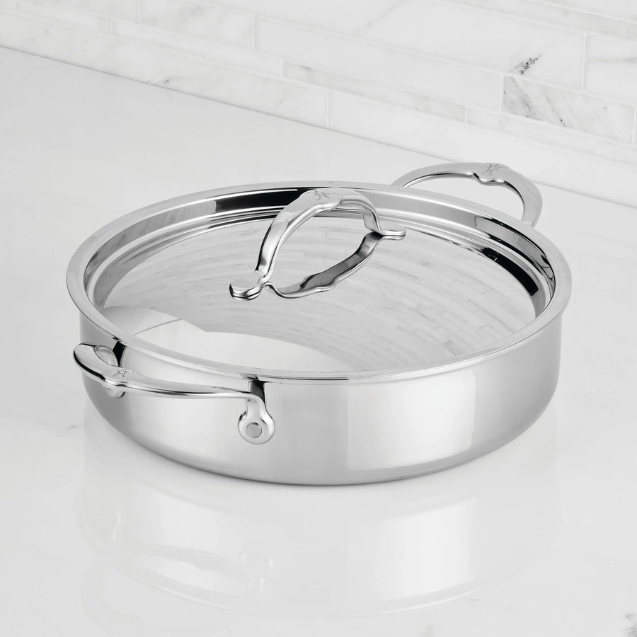A ProBond® Luxe™ Polished Clad Stainless Steel Rondeau with lid and dual handles rests on a white marble countertop, set against a backdrop of white tile backsplash.