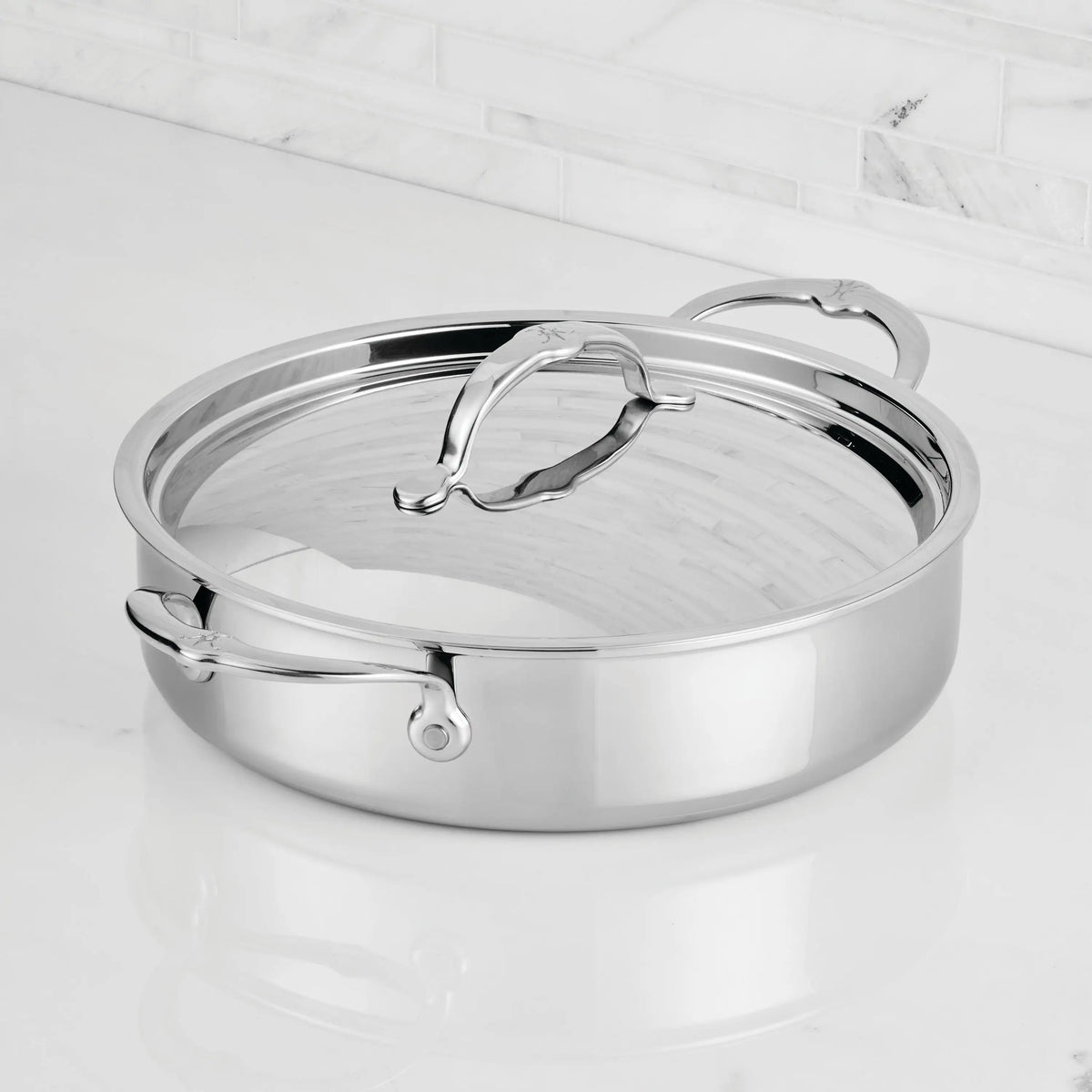 A ProBond® Luxe™ Polished Clad Stainless Steel Rondeau with lid and dual handles rests on a white marble countertop, set against a backdrop of white tile backsplash.