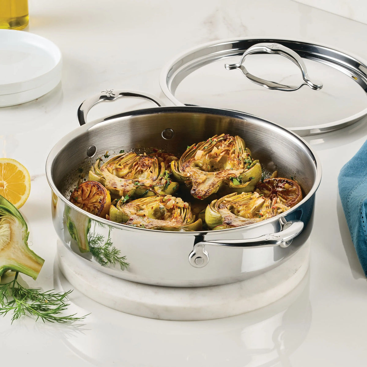 A ProBond® Luxe™ Polished Clad Stainless Steel Rondeau with lid sits on marble, holding browned halved artichokes. Fresh herbs, a lemon, and a blue napkin are arranged nearby.