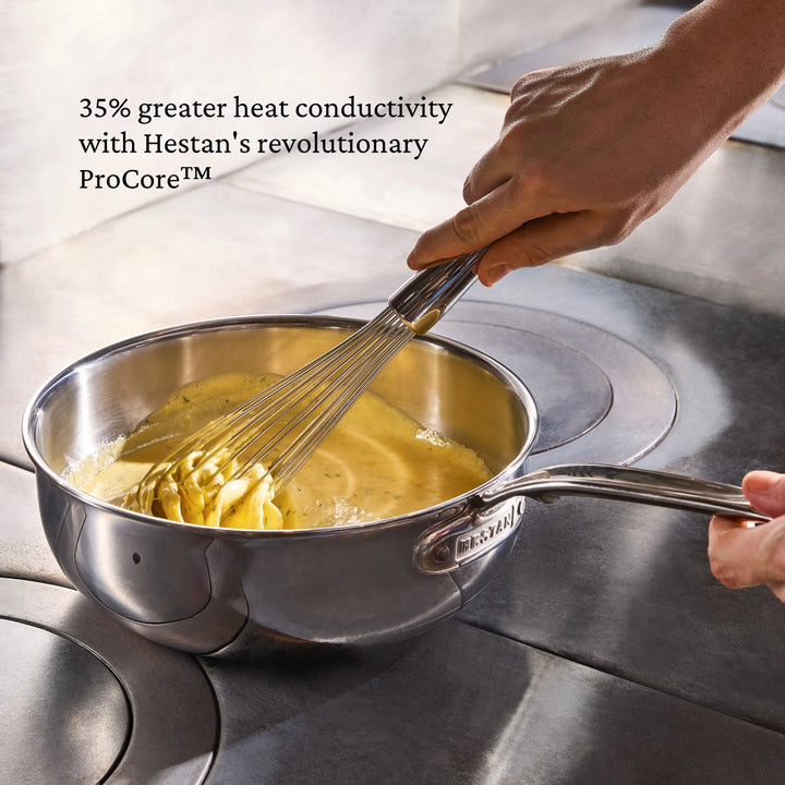 A person whisks a creamy yellow mixture in a ProBond® Luxe™ Professional Clad Stainless Steel Essential Pan on the stove. Text: “ProCore™ delivers 35% greater heat conductivity—enjoy professional results with ProBond® Luxe™ cookware.”.