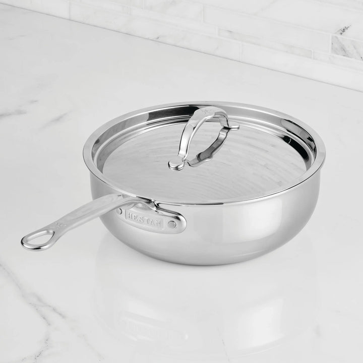 The ProBond® Luxe™ Professional Clad Stainless Steel Essential Pan with Lid features a sleek, reflective finish and long handle, resting securely on a white marble countertop.