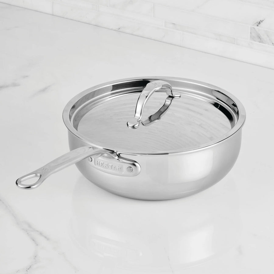 The ProBond® Luxe™ Professional Clad Stainless Steel Essential Pan with Lid features a sleek, reflective finish and long handle, resting securely on a white marble countertop.
