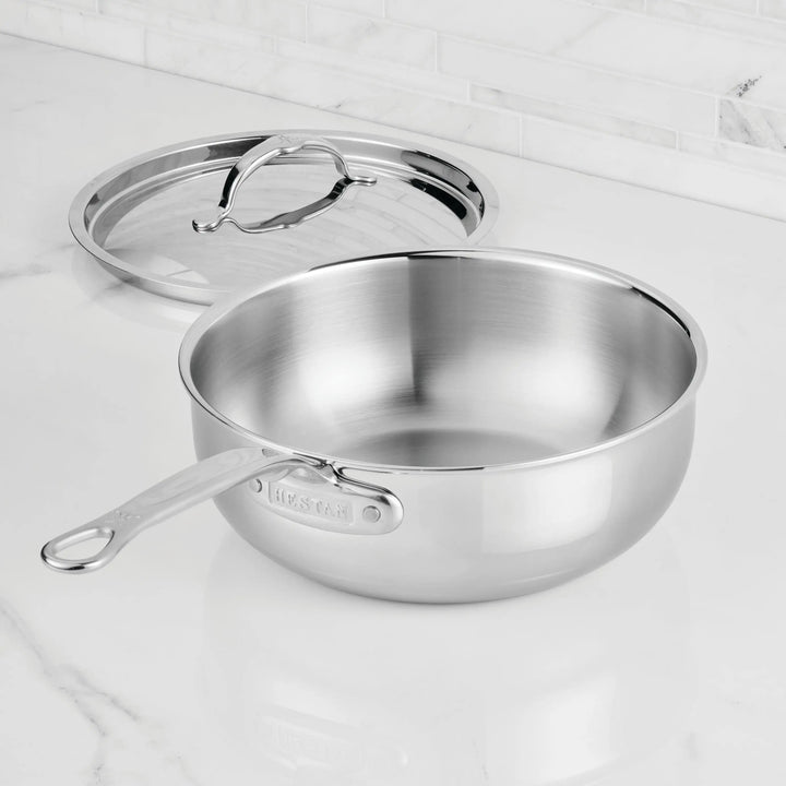 A ProBond® Luxe™ Professional Clad Stainless Steel Essential Pan with Lid sits on a white marble countertop, highlighting its Italian craftsmanship and modern design.