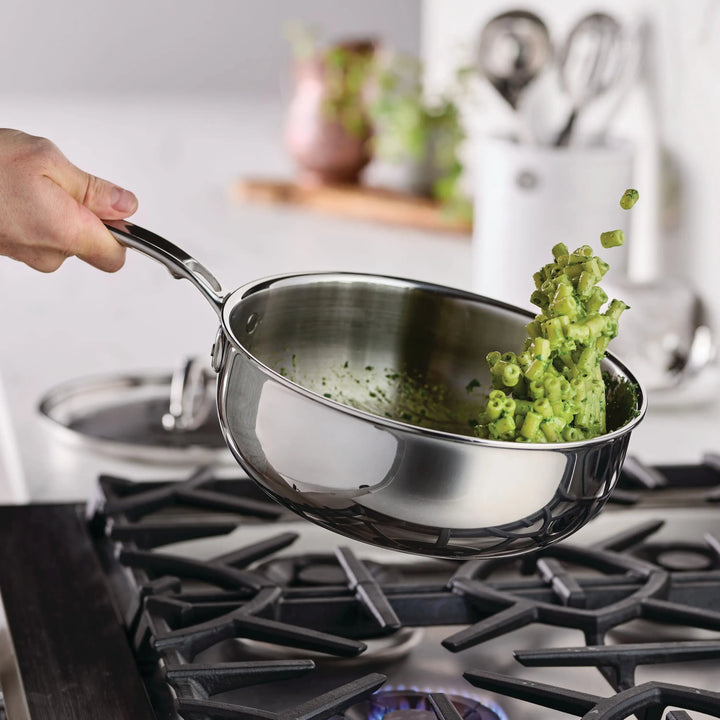 A hand holds a ProBond® Luxe™ Professional Clad Stainless Steel Essential Pan with Lid over a gas stove, flipping chopped green vegetables in mid-air. A glass lid rests nearby on the countertop, with a softly blurred kitchen background.